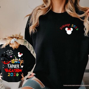 Puede incluir: Una sudadera negra de manga larga con un peque&ntilde;o icono blanco de la cabeza de Mickey Mouse con un lazo rojo, el texto DISNEY 2026 y un gr&aacute;fico grande en la espalda con personajes de Disney, fuegos artificiales y el texto DISNEY FAMILY VACATION 2026.