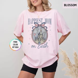 May include: A light pink t-shirt with the words "Happiest Mom on Earth" in a stylized font. The design includes a castle illustration within a decorative oval frame, with a bow and balloons. The shirt is a relaxed fit.