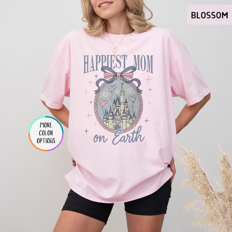 May include: A light pink t-shirt with the words "Happiest Mom on Earth" in a stylized font. The design includes a castle illustration within a decorative oval frame, with a bow and balloons. The shirt is a relaxed fit.