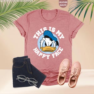 Op de afbeelding: Heather mauve t-shirt met een afbeelding van Donald Duck en de tekst "THIS IS MY HAPPY FACE". De afbeelding toont ook een donkere jeans, een bril met helder montuur en roze sneakers.