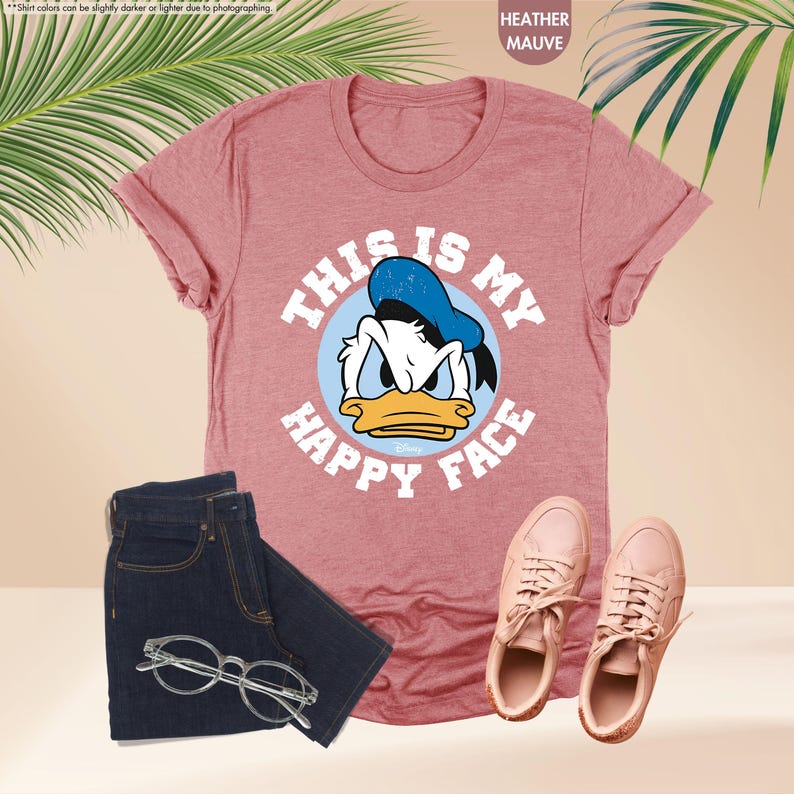 Op de afbeelding: Heather mauve t-shirt met een afbeelding van Donald Duck en de tekst "THIS IS MY HAPPY FACE". De afbeelding toont ook een donkere jeans, een bril met helder montuur en roze sneakers.