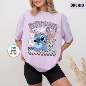 May include: Orchid-colored t-shirt featuring a graphic of Stitch, ducks, and flowers. The word "STITCH" is arched across the top. The design includes a checkerboard pattern at the bottom. The shirt has short sleeves.