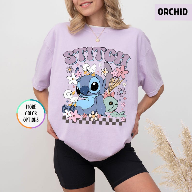 May include: Orchid-colored t-shirt featuring a graphic of Stitch, ducks, and flowers. The word "STITCH" is arched across the top. The design includes a checkerboard pattern at the bottom. The shirt has short sleeves.