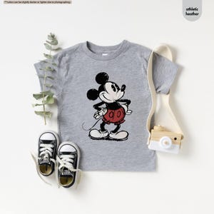 May include: A gray athletic heather t-shirt featuring a distressed graphic of Mickey Mouse. Mickey is depicted in black, red, and white, with his hands on his hips. The shirt is paired with black canvas sneakers.