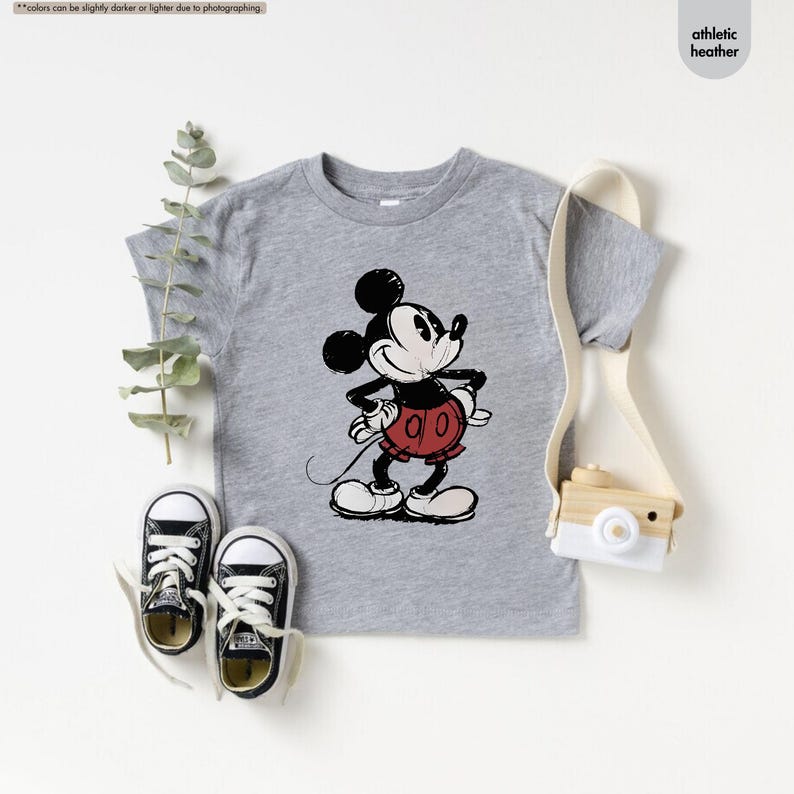 May include: A gray athletic heather t-shirt featuring a distressed graphic of Mickey Mouse. Mickey is depicted in black, red, and white, with his hands on his hips. The shirt is paired with black canvas sneakers.