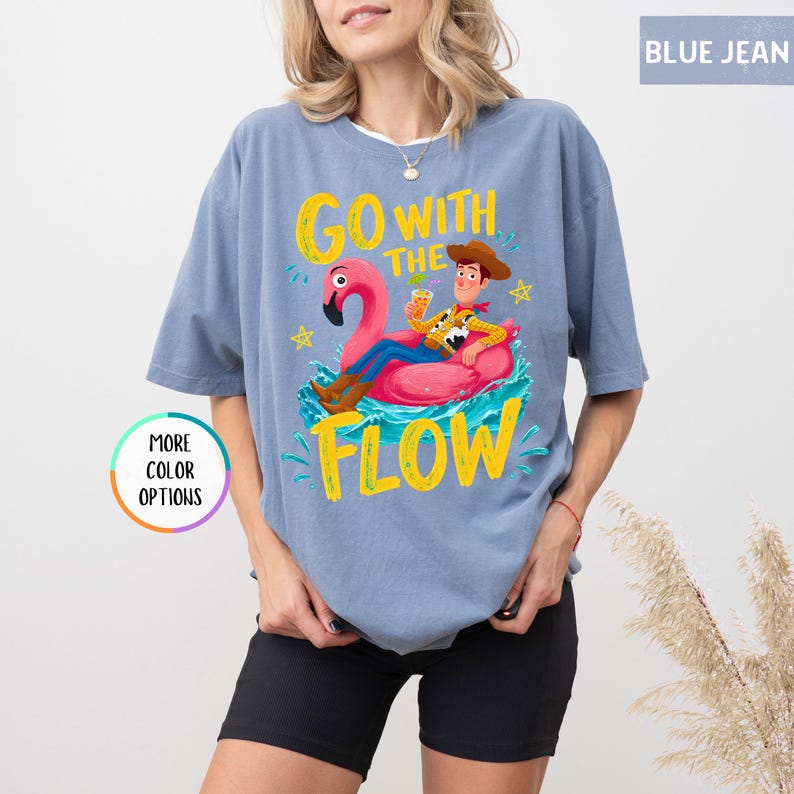 Puede incluir: Una camiseta color azul jean con un gr&aacute;fico de un vaquero de dibujos animados sentado en un flotador de flamenco rosa en agua azul. El texto GO WITH THE FLOW est&aacute; impreso en amarillo sobre y bajo el gr&aacute;fico. Un icono circular dice MORE COLOR OPTIONS.