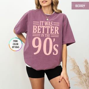 May include: A berry-colored short-sleeve t-shirt featuring the text IT WAS BETTER IN THE 90's printed in a light pink, distressed font. The shirt has a crew neckline and a relaxed fit, with a small circular graphic indicating more color options.