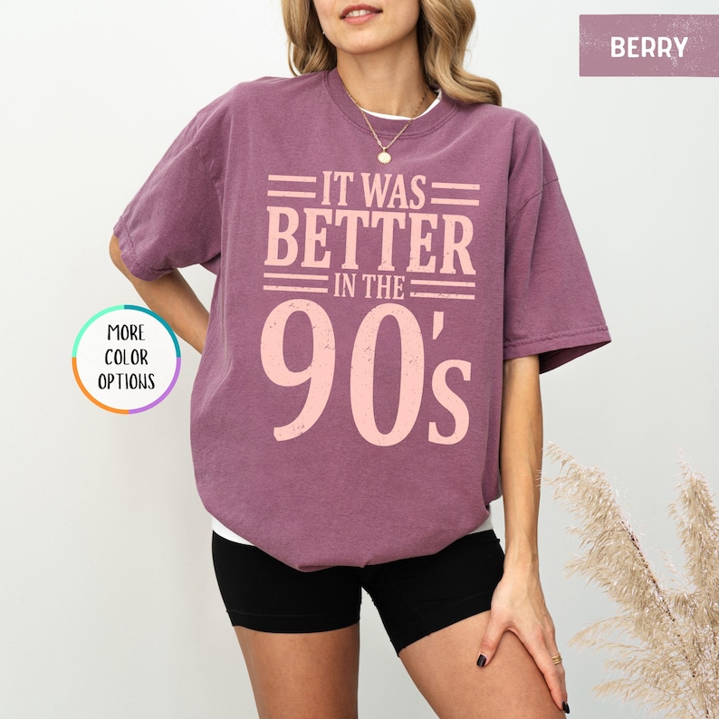 May include: A berry-colored short-sleeve t-shirt featuring the text IT WAS BETTER IN THE 90's printed in a light pink, distressed font. The shirt has a crew neckline and a relaxed fit, with a small circular graphic indicating more color options.