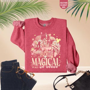 May include: Heather scarlet red sweatshirt with a graphic design featuring a castle, Mickey Mouse ears, and the text "The Most Magical Place". The sweatshirt is paired with blue jeans, glasses, and a black purse.