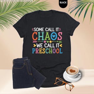 May include: Black t-shirt with the text "SOME CALL IT CHAOS WE CALL IT PRESCHOOL" in colorful letters. The shirt is paired with dark blue jeans, a cup of coffee with a heart design, and orange eyeglasses.