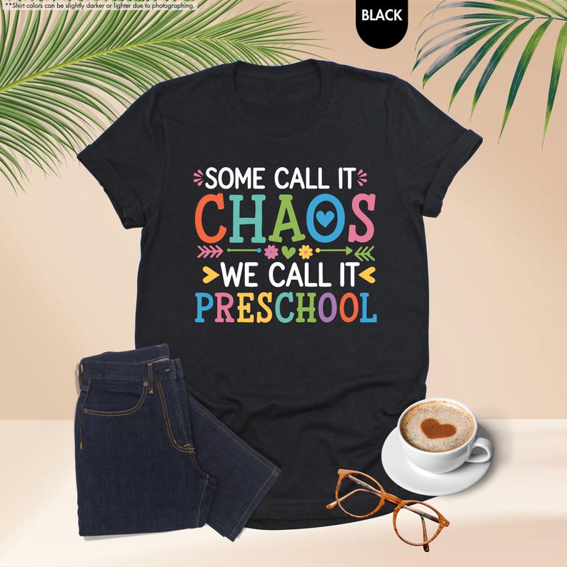 May include: Black t-shirt with the text "SOME CALL IT CHAOS WE CALL IT PRESCHOOL" in colorful letters. The shirt is paired with dark blue jeans, a cup of coffee with a heart design, and orange eyeglasses.