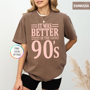 May include: A brown short-sleeve crew neck t-shirt featuring the light pink text "IT WAS BETTER IN THE 90's" printed on the front. The shirt has a relaxed fit and a solid color design. A small circular graphic on the left reads "MORE COLOR OPTIONS".