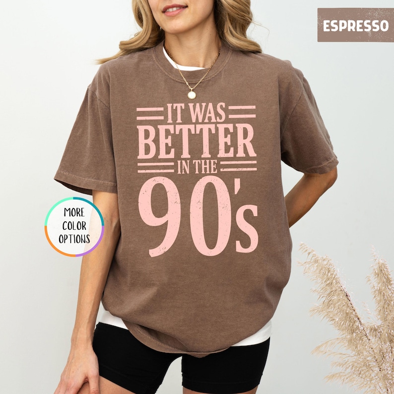 May include: A brown short-sleeve crew neck t-shirt featuring the light pink text "IT WAS BETTER IN THE 90's" printed on the front. The shirt has a relaxed fit and a solid color design. A small circular graphic on the left reads "MORE COLOR OPTIONS".