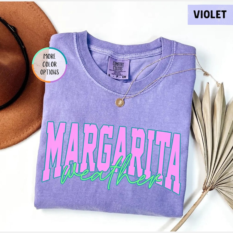 May include: A violet-colored t-shirt with the words "MARGARITA weather" in a pink and green font. The shirt has a crew neck and short sleeves. A gold necklace with a pendant is draped on the shirt. The words "MORE COLOR OPTIONS" are in a circle on the left.