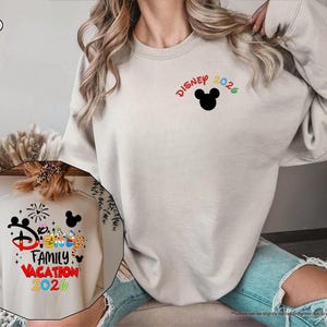 Puede incluir: Una sudadera de cuello redondo color arena que presenta una peque&ntilde;a silueta negra de Mickey Mouse con el texto Disney 2026 en el frente. El dise&ntilde;o posterior muestra el texto Disney Family Vacation 2026 con gr&aacute;ficos de personajes coloridos y fuegos artificiales.