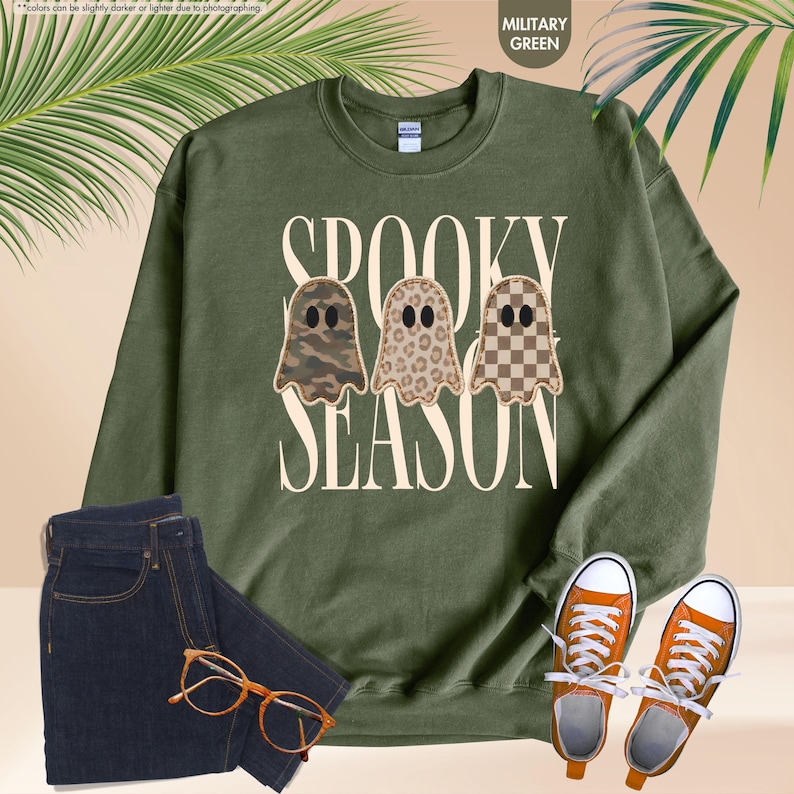 May include: A military green crewneck sweatshirt featuring the text SPOOKY SEASON in cream lettering. The design includes three ghost illustrations with different patterns: camouflage, leopard print, and a brown and cream checkerboard pattern.