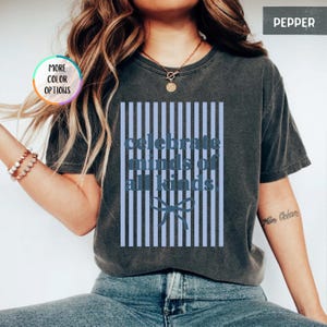 Puede incluir: Camiseta gris oscuro con el texto azul "celebrate minds of all kinds". El texto est&aacute; sobre un fondo a rayas azul claro con un lazo. La camiseta tiene mangas cortas y cuello redondo.