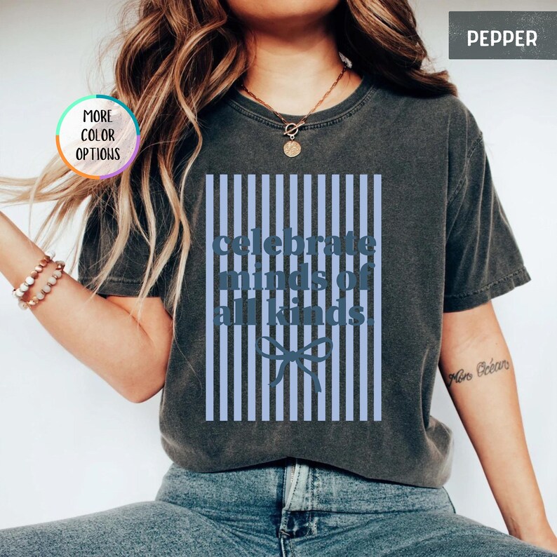 Puede incluir: Camiseta gris oscuro con el texto azul "celebrate minds of all kinds". El texto est&aacute; sobre un fondo a rayas azul claro con un lazo. La camiseta tiene mangas cortas y cuello redondo.