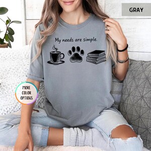 May include: Gray t-shirt with the phrase "My needs are simple." The graphic includes a coffee cup, a paw print, and a stack of books. The shirt is a solid gray color.