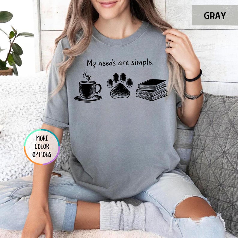 May include: Gray t-shirt with the phrase "My needs are simple." The graphic includes a coffee cup, a paw print, and a stack of books. The shirt is a solid gray color.