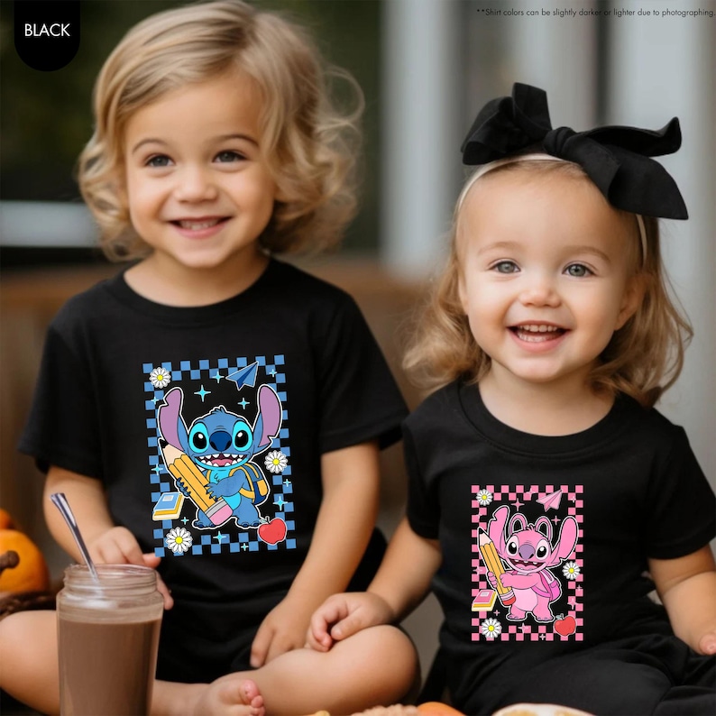 May include: Two children wearing black t-shirts with cartoon character graphics. One shirt features a blue character with a pencil, backpack, and apple. The other shirt has a pink character with a pencil and apple.