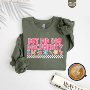 Puede incluir: Sudadera verde militar con el texto "BUT DID YOU DOCUMENT?" en rosa y blanco. El dise&ntilde;o incluye gr&aacute;ficos de tem&aacute;tica m&eacute;dica como una venda, un estetoscopio y una jeringa. Un patr&oacute;n de cuadros blancos y negros en la parte inferior.