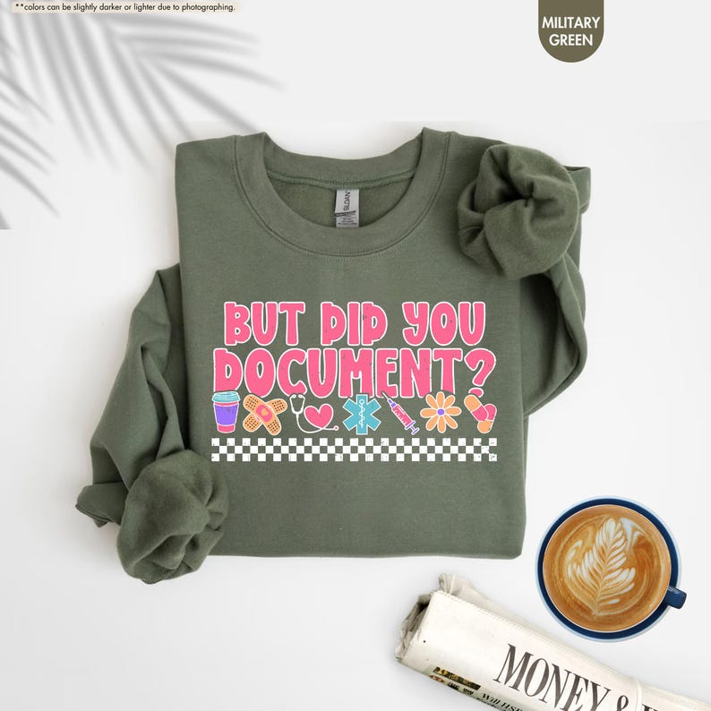 Puede incluir: Sudadera verde militar con el texto "BUT DID YOU DOCUMENT?" en rosa y blanco. El dise&ntilde;o incluye gr&aacute;ficos de tem&aacute;tica m&eacute;dica como una venda, un estetoscopio y una jeringa. Un patr&oacute;n de cuadros blancos y negros en la parte inferior.
