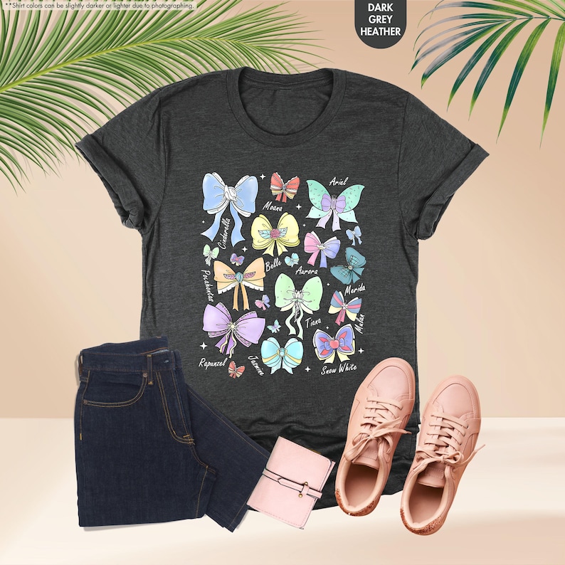 May include: Dark grey heather t-shirt with a colourful bow graphic. The graphic features various bows in pastel colours with names like Ariel, Belle, and Rapunzel. The image also includes dark wash jeans, a pink wallet, and pink trainers.