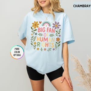 May include: Light blue t-shirt with the text "BIG FAN OF HUMAN RIGHTS" in colorful letters, surrounded by floral and rainbow graphics. The shirt has short sleeves and a crew neck.