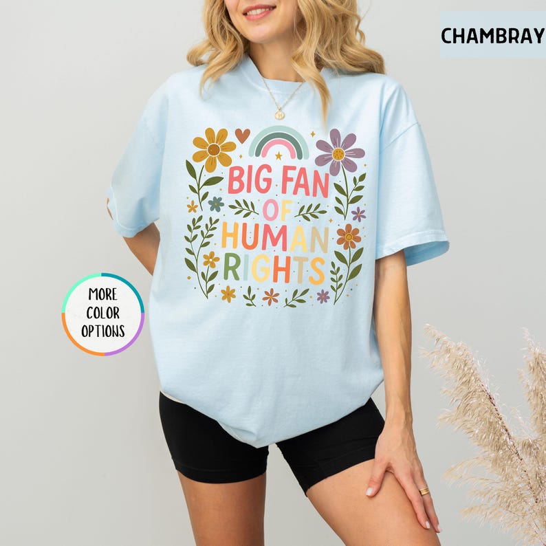 May include: Light blue t-shirt with the text "BIG FAN OF HUMAN RIGHTS" in colorful letters, surrounded by floral and rainbow graphics. The shirt has short sleeves and a crew neck.