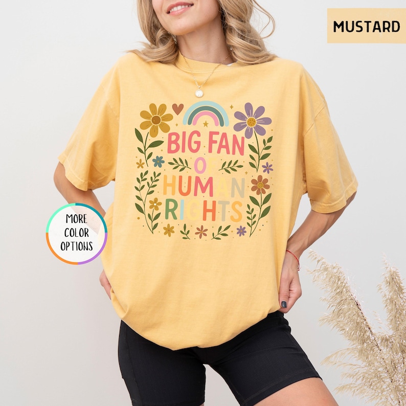 May include: Mustard-colored t-shirt with a floral design and the text "BIG FAN OF HUMAN RIGHTS." The design includes a rainbow, flowers, and leaves. The shirt has short sleeves and a crew neck.