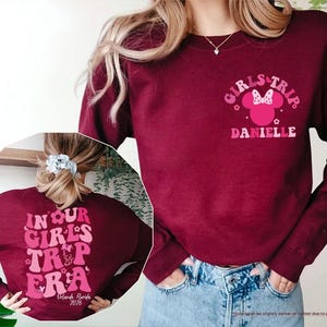 Peut inclure: Sweat-shirt bordeaux avec l'inscription "GIRLS TRIP DANIELLE" sur le devant et "IN OUR GIRLS TRIP ERA Orlando, Florida 2026" au dos. Le motif comprend une silhouette de Minnie Mouse et des &eacute;l&eacute;ments floraux. Le sweat-shirt est &agrave; manches longues et col rond.