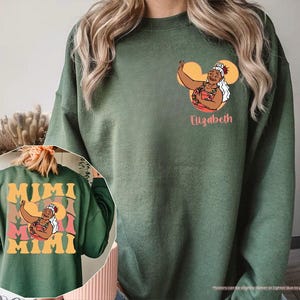 May include: Military green sweatshirt with a graphic of a woman in a red and pink floral top, with the name "Elizabeth" below. The back of the sweatshirt has the word "MIMI" in large, retro-style lettering, with the same graphic.