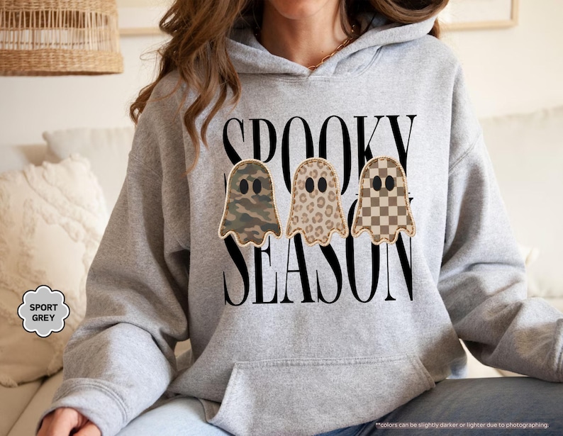 May include: A sport grey hooded sweatshirt featuring the black text SPOOKY SEASON and three ghost illustrations. The ghosts have distinct patterns: one with a green camouflage print, one with a brown leopard print, and one with a tan checkerboard pattern.
