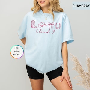 May include: Light blue t-shirt with pink cowboy-themed graphics and the words "Cloud 9". The design includes a cowboy boot, hat, bow, and horseshoe. The shirt is a relaxed fit.