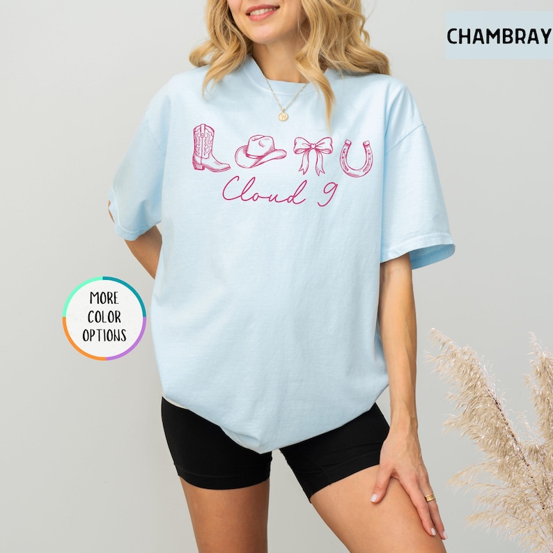 May include: Light blue t-shirt with pink cowboy-themed graphics and the words "Cloud 9". The design includes a cowboy boot, hat, bow, and horseshoe. The shirt is a relaxed fit.