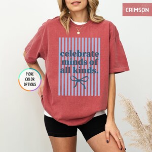 Puede incluir: Camiseta color carmes&iacute; con el texto "celebrate minds of all kinds". El texto est&aacute; dentro de un rect&aacute;ngulo a rayas azules y rojas con un lazo azul en la parte inferior. La camiseta tiene mangas cortas y un corte holgado.
