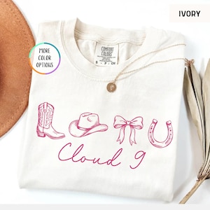 May include: Ivory-colored t-shirt with a pink graphic of a cowboy boot, hat, bow, and horseshoe, with the words "Cloud 9" in cursive. The shirt has a crew neck and short sleeves.