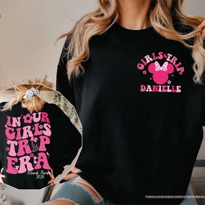 Peut inclure: Sweat-shirt noir avec texte et graphiques roses. Le devant affiche "GIRLS TRIP" avec une t&ecirc;te de Minnie Mouse et le nom "DANIELLE". Le dos indique "IN OUR GIRLS TRIP ERA" avec "Orlando, Florida 2026".