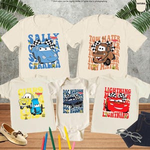 May include: Five off-white t-shirts and a onesie featuring cartoon car characters. The shirts display characters like Sally Carrera, Tow Mater, and Lightning McQueen. The onesie features Doc Hudson. A pair of shoes, jeans, and glasses are also in the image.