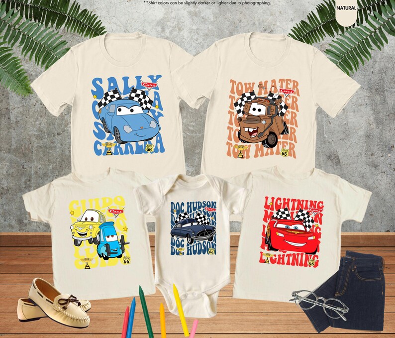 May include: Five off-white t-shirts and a onesie featuring cartoon car characters. The shirts display characters like Sally Carrera, Tow Mater, and Lightning McQueen. The onesie features Doc Hudson. A pair of shoes, jeans, and glasses are also in the image.