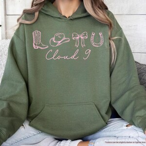May include: Military green hoodie with a front pocket and a drawstring hood. The front features pink line art of a cowboy boot, hat, bow, horseshoe, and the words "Cloud 9".