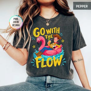 Puede incluir: Una camiseta gris carb&oacute;n con un gr&aacute;fico de un personaje vaquero sentado en un flotador de flamenco rosa sobre agua azul. El texto "GO WITH THE FLOW" aparece en letras amarillas. La camiseta incluye una insignia "MORE COLOR OPTIONS" y una etiqueta "PEPPER".