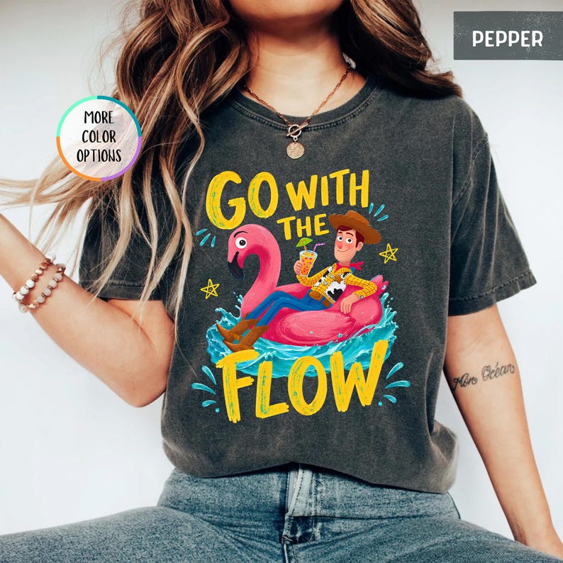 Puede incluir: Una camiseta gris carb&oacute;n con un gr&aacute;fico de un personaje vaquero sentado en un flotador de flamenco rosa sobre agua azul. El texto "GO WITH THE FLOW" aparece en letras amarillas. La camiseta incluye una insignia "MORE COLOR OPTIONS" y una etiqueta "PEPPER".