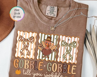 May include: Espresso-colored t-shirt with a Thanksgiving-themed graphic. The design features a turkey wearing a pilgrim hat, bows, and the text "GOBBLE + GOBBLE till you wobble". A silver necklace is draped across the shirt.