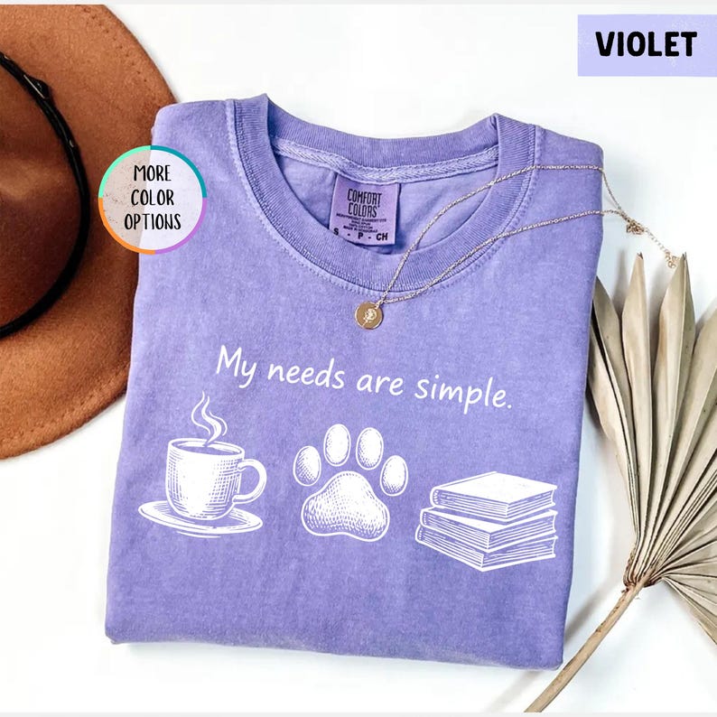 May include: A violet Comfort Colors t-shirt with the phrase "My needs are simple" printed in white. The design includes a coffee cup, paw print, and books. A gold necklace rests on the shirt.