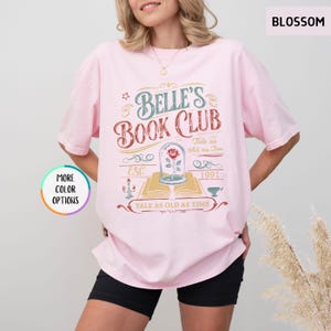 Pu&ograve; includere: T-shirt rosa chiaro con la scritta "Belle's Book Club" in un design vintage. La grafica include una rosa in una cupola di vetro, la frase "Tale as old as Time" e l'anno "1991". La maglietta ha anche la parola "Blossom".