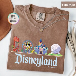 Op de afbeelding: Espresso-kleurig T-shirt met een Disneyland Resort-afbeelding. Het ontwerp bevat kleurrijke illustraties van iconische parklandmarks, waaronder een kasteel, een reuzenrad met Mickey Mouse en een bol. Een zilveren ketting met een hanger is over het shirt gedrapeerd.