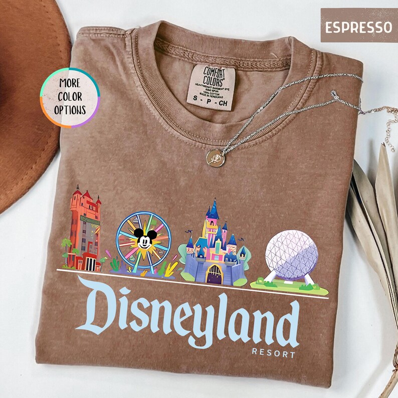 Op de afbeelding: Espresso-kleurig T-shirt met een Disneyland Resort-afbeelding. Het ontwerp bevat kleurrijke illustraties van iconische parklandmarks, waaronder een kasteel, een reuzenrad met Mickey Mouse en een bol. Een zilveren ketting met een hanger is over het shirt gedrapeerd.
