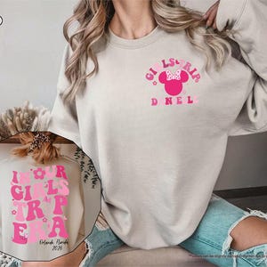 Peut inclure: Un sweat-shirt couleur sable avec l'inscription "GIRLS TRIP" en rose, orn&eacute; d'un motif de t&ecirc;te de Minnie Mouse. Le sweat-shirt porte &eacute;galement l'inscription "IN OUR GIRLS TRIP ERA" et "Orlando, Florida 2026" dans une image circulaire.